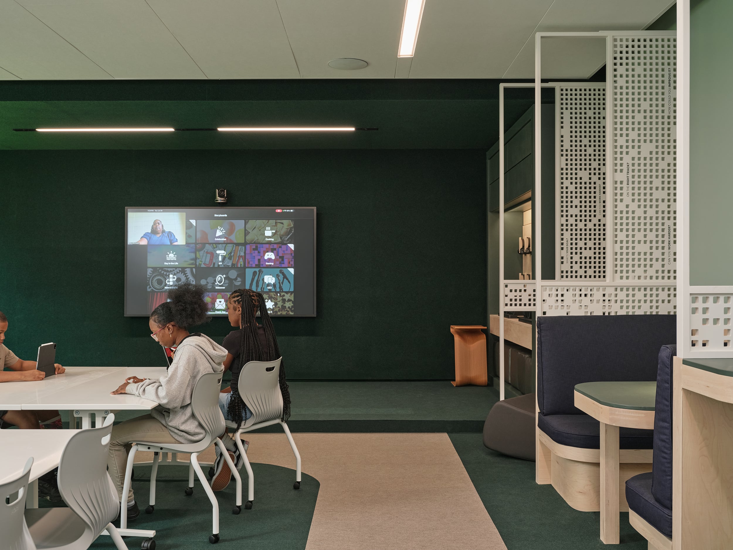 Several students sit at tables using tablets in a modern classroom, while a large screen displays a video call with multiple people.