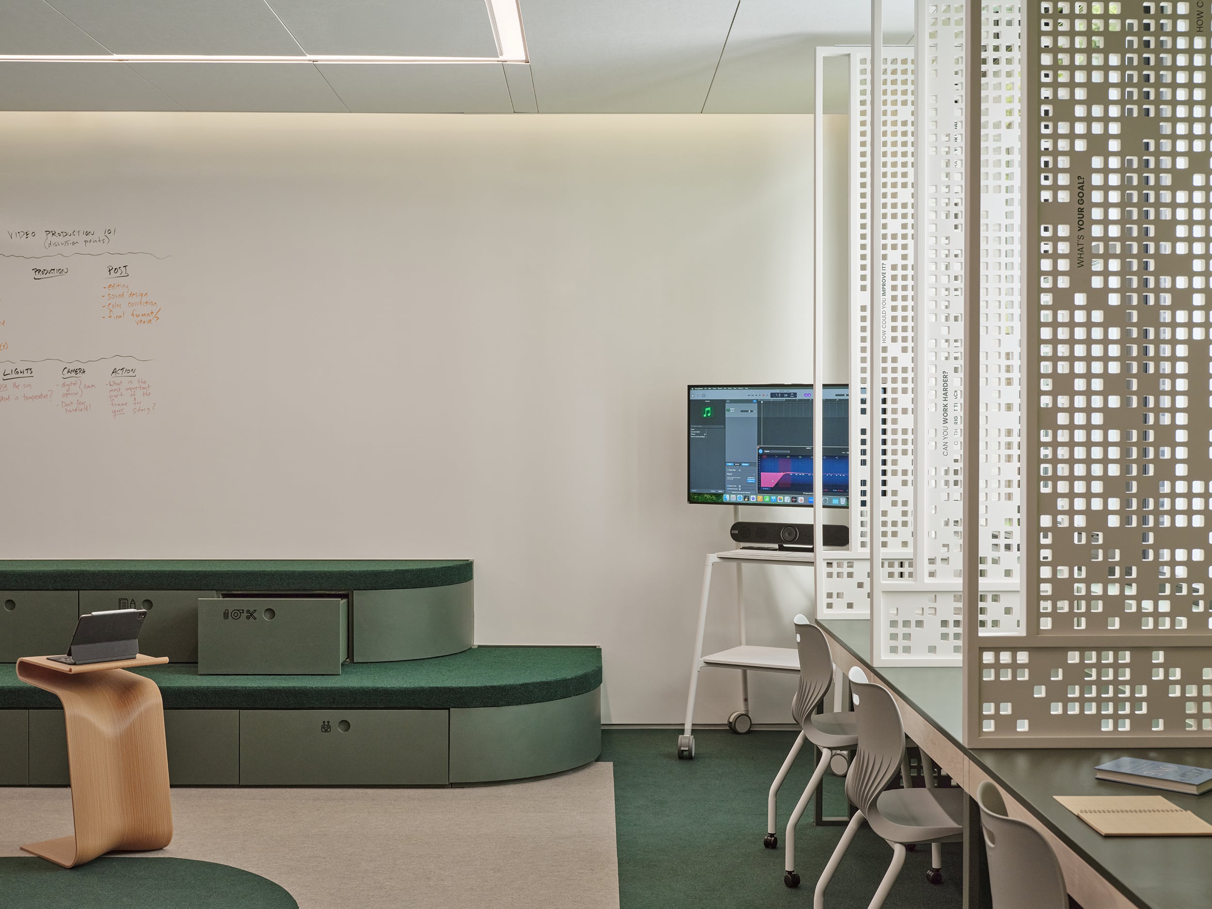 Modern office space with tiered green seating, a small wooden table, a TV monitor on a stand, whiteboard with notes, and a perforated partition next to workstations with chairs.