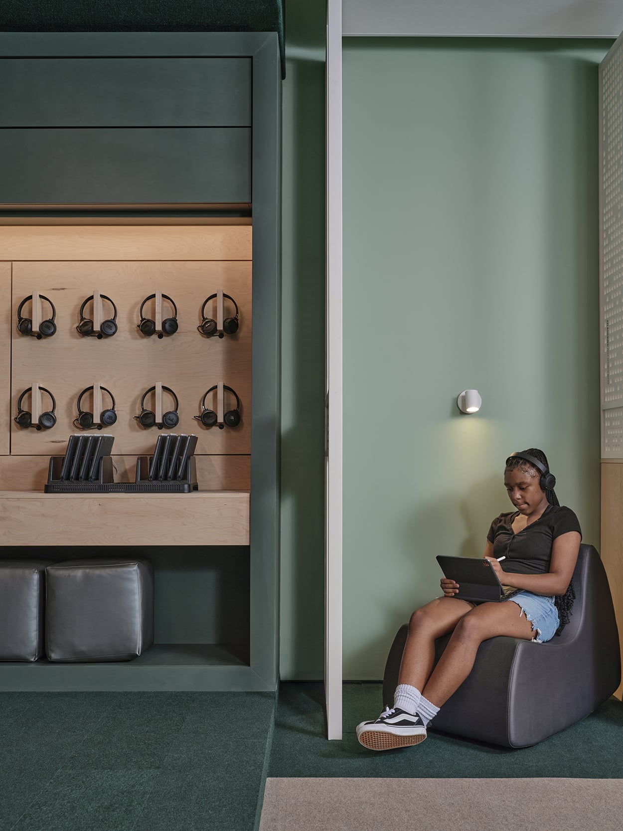 A person sits on a chair wearing headphones and using a tablet in a modern room; several pairs of headphones hang on the wall nearby.