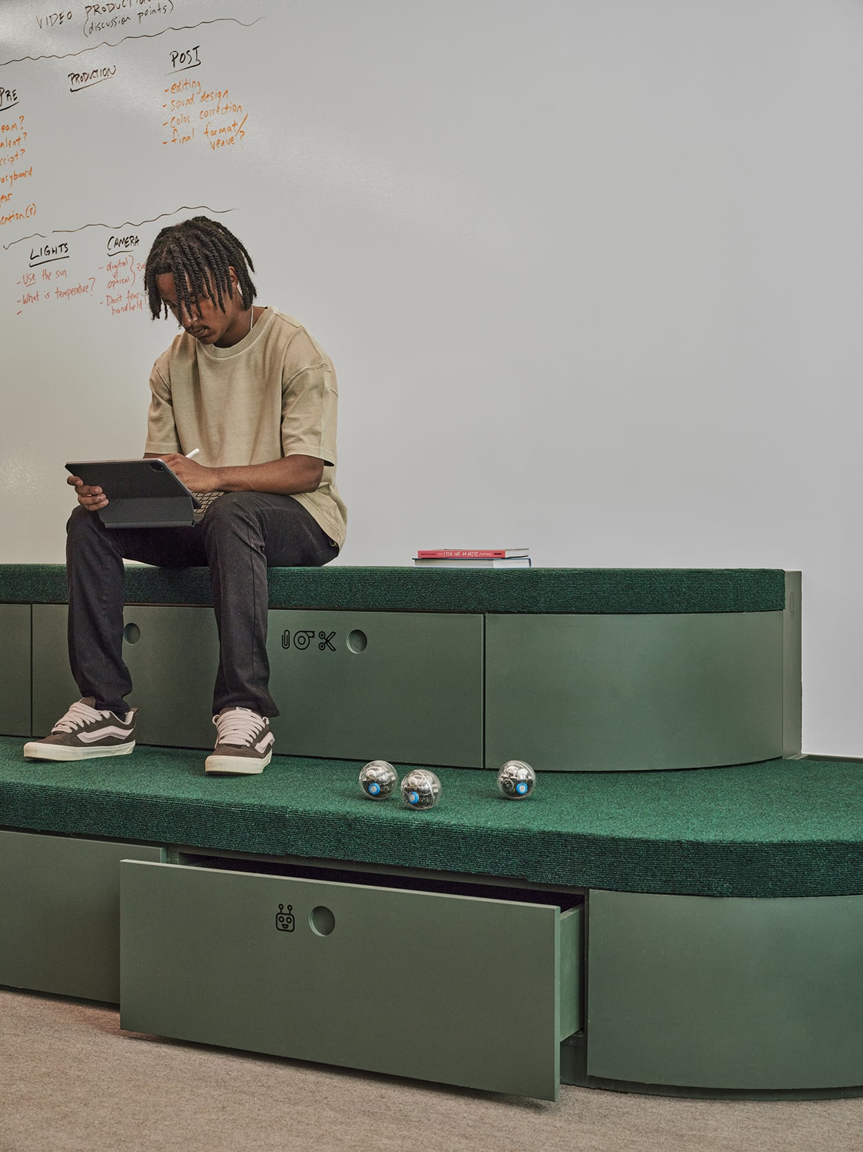 A person sits on green tiered seating, using a tablet. Three small spherical robots are on the steps. A whiteboard with notes is visible in the background.