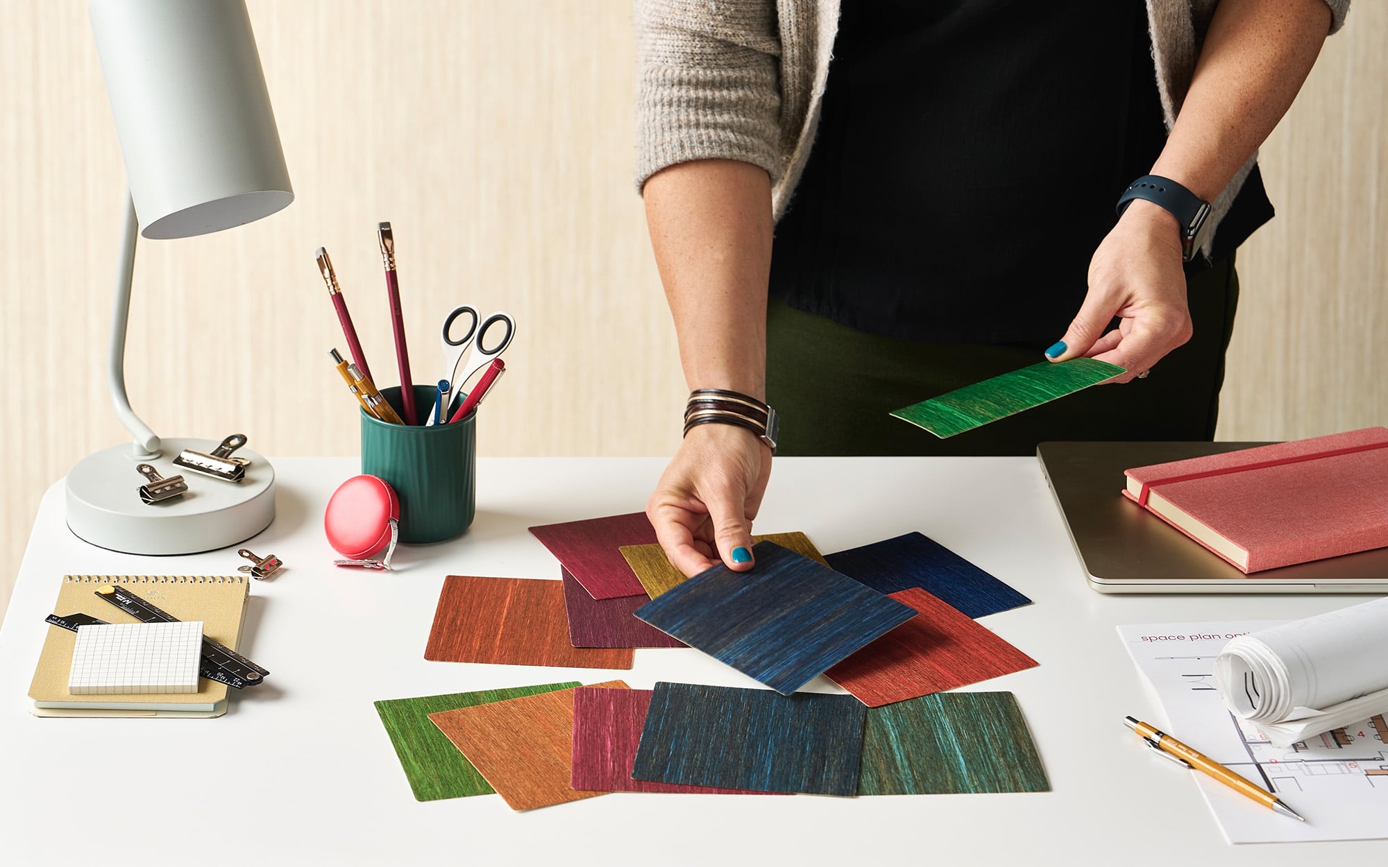 A person stands at a desk in the Metropolis Sustainability Lab, sorting through colorful rectangular material samples, surrounded by office supplies, a lamp, and some paperwork.