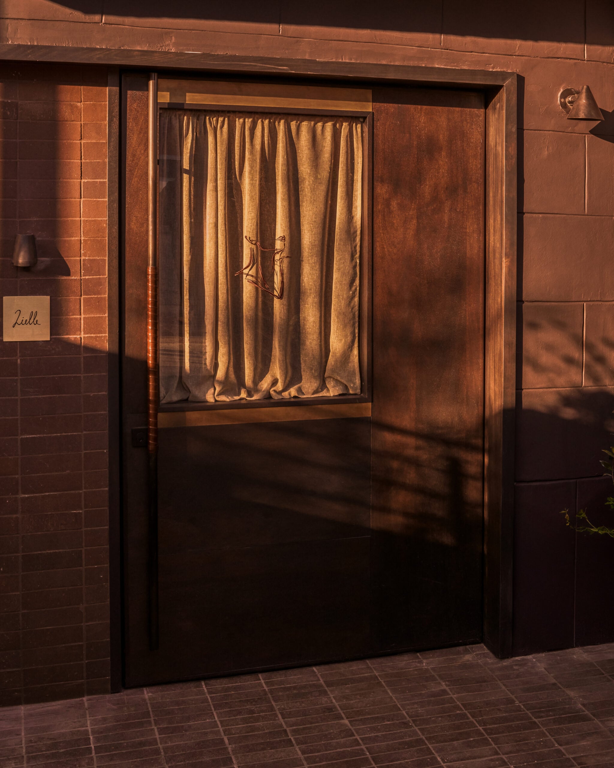 A brown wooden door with a light brown curtain covering a window, illuminated by warm sunlight. A small sign reading "Luli" is mounted on the wall to the left, marking a quiet place where lovers unite.