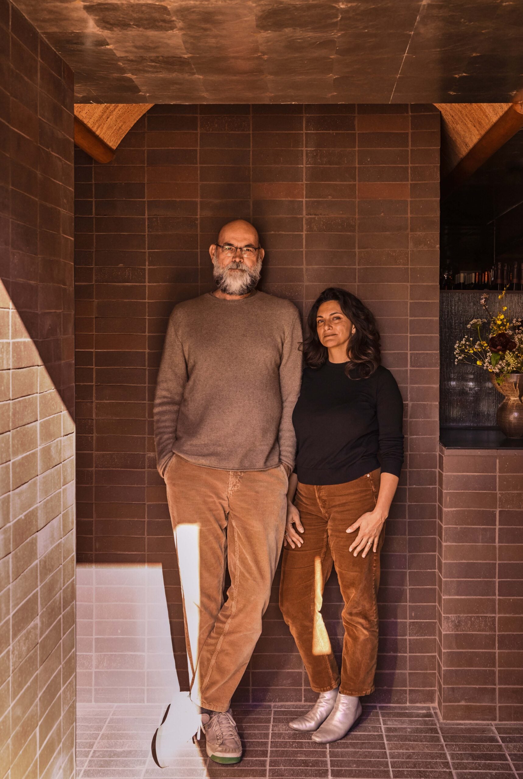 A man and a woman stand side by side against a tiled brown wall, both wearing brown pants and neutral tops, with natural light illuminating part of the space.