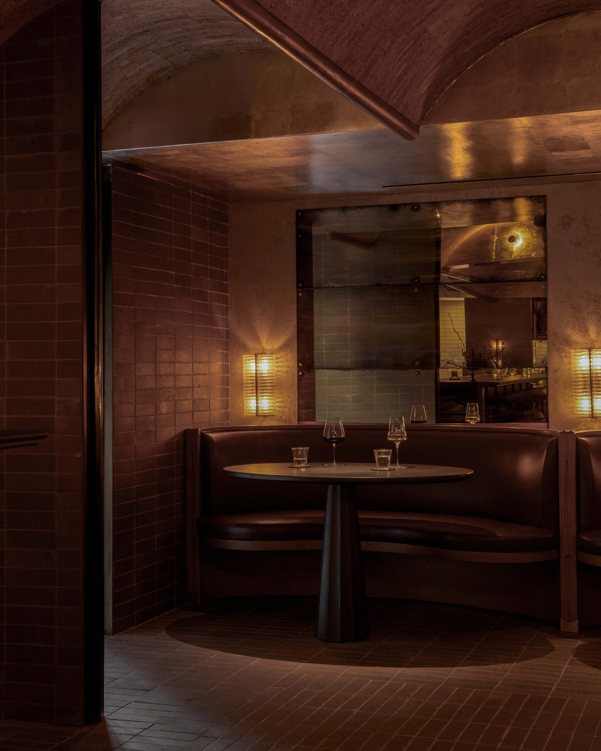 A dimly lit restaurant booth with a round table, brown leather seating, and several empty glasses; warm lighting and tiled walls create a cozy atmosphere where lovers unite.