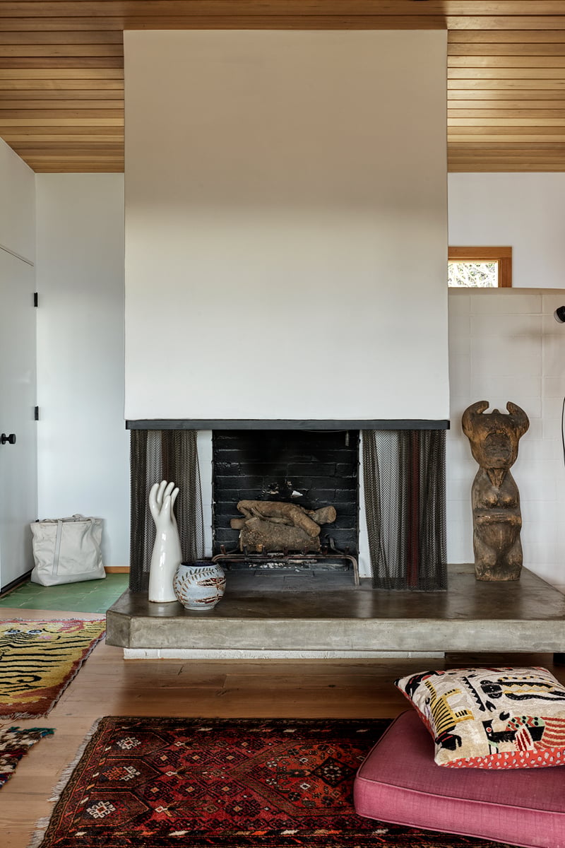 A modern fireplace with a white mantel and dark interior is flanked by ceramic vases, a sculptural wooden figure, and patterned rugs and cushions, creating a cozy spot where lovers unite in warmth and style.