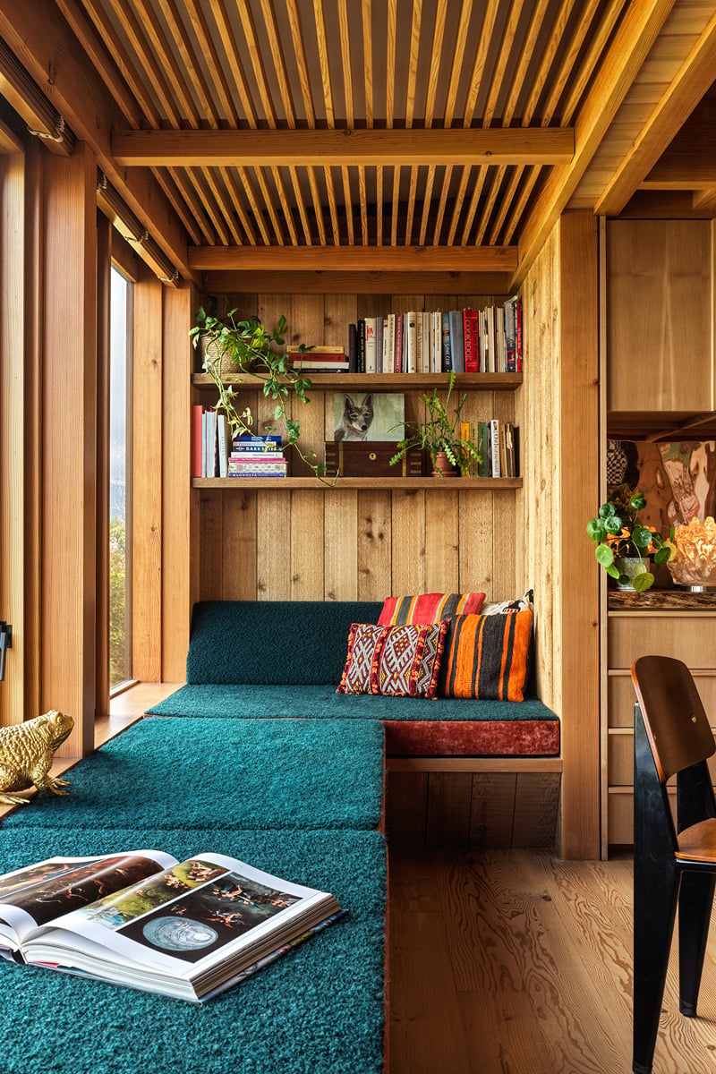 A cozy reading nook with teal cushions, colorful pillows, wooden shelves with books and plants, and natural wood paneling, next to a large window. An open book rests on the seat where lovers unite over shared stories.