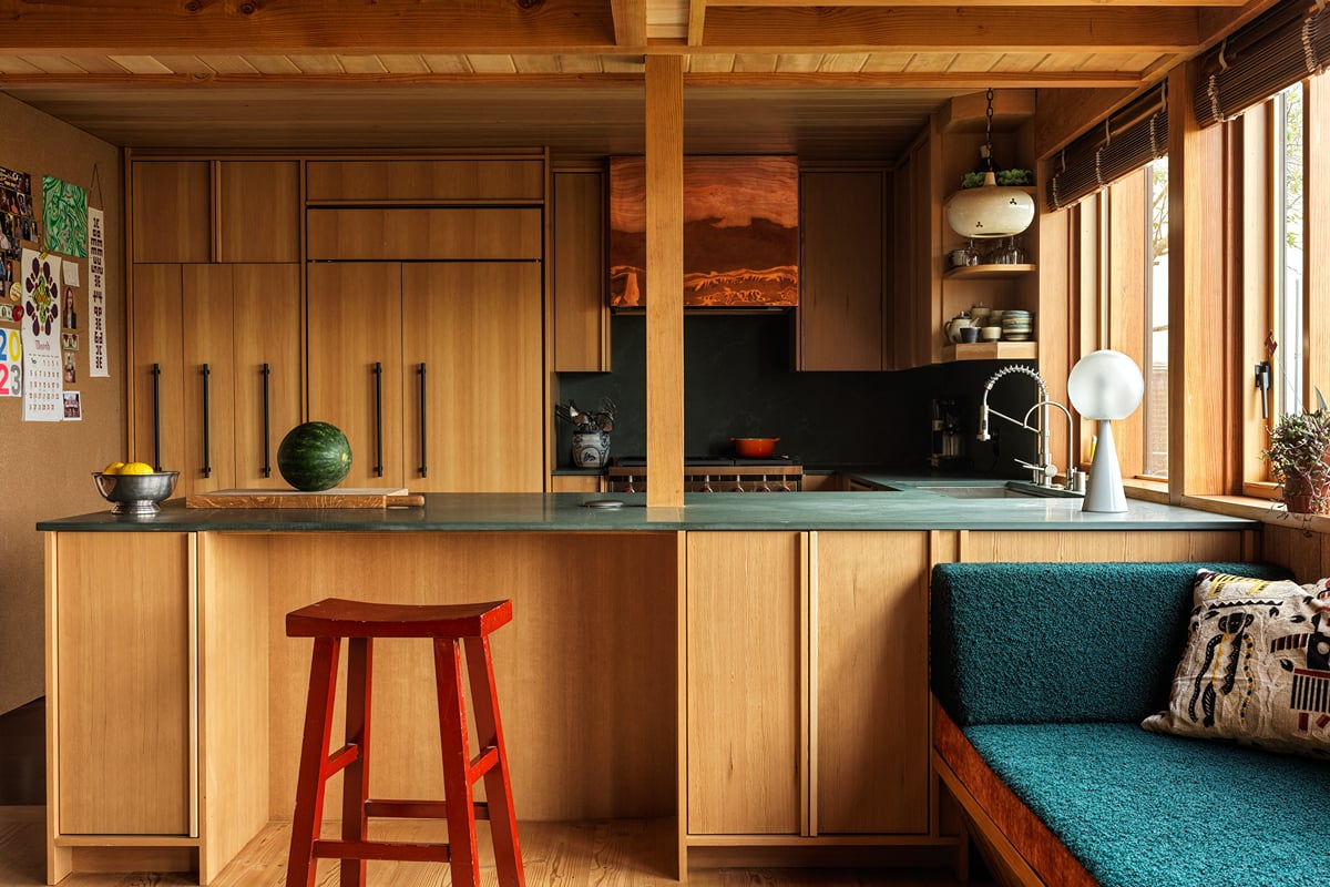 Modern kitchen with wood cabinets, green countertop, red stool, and a watermelon on the counter—where lovers unite over shared meals by the window with blinds and a built-in bench with teal upholstery and patterned pillows.