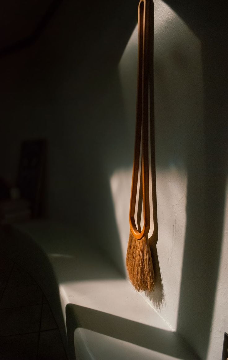 A hand broom with a long looped handle, crafted with fine textiles, hangs on a white wall, illuminated by sunlight casting distinct shadows.