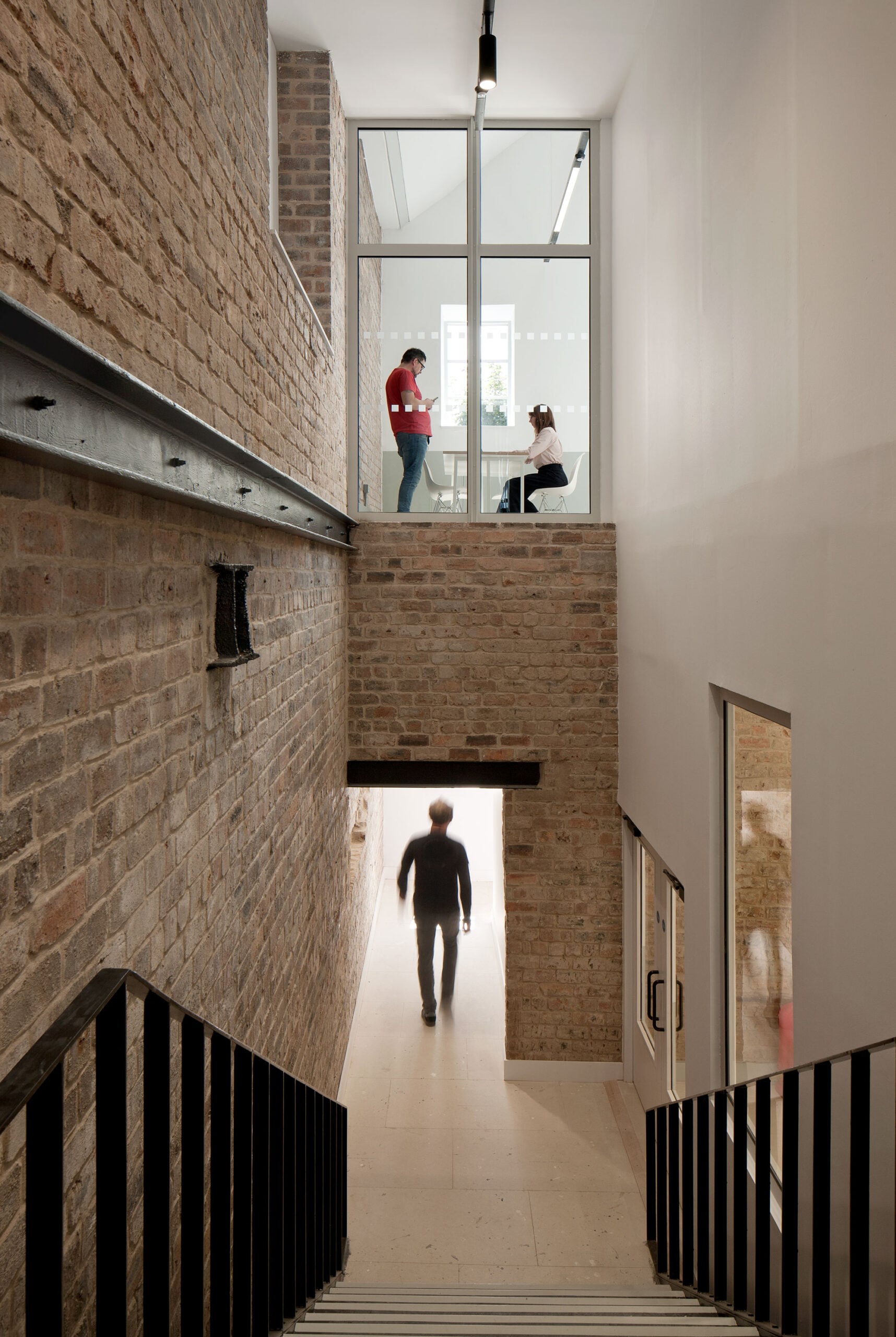 A person walks down a stairwell with exposed brick walls, while two people converse in a glass-walled room above.