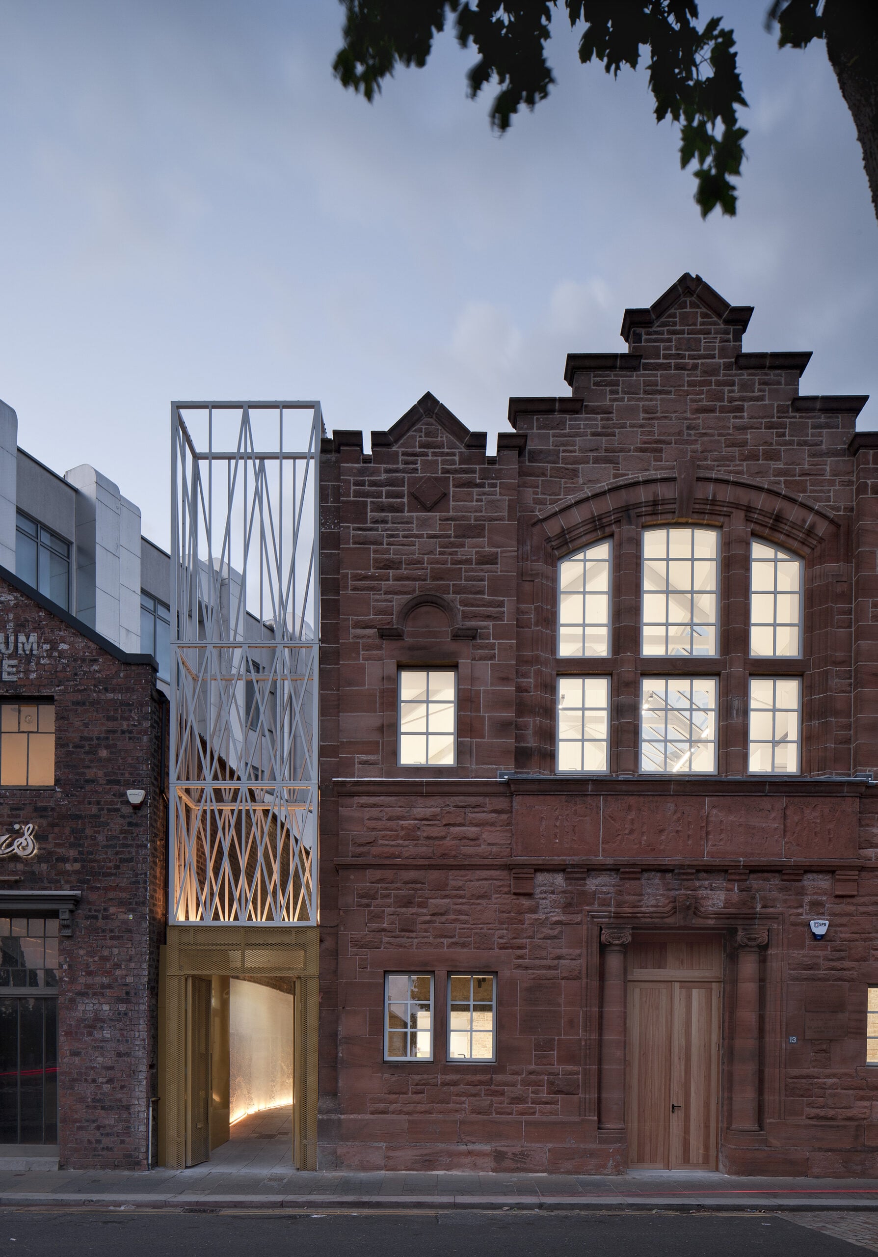 A modern, gold-accented metal and glass extension contrasts with the adjoining historic red brick building on a city street at dusk.