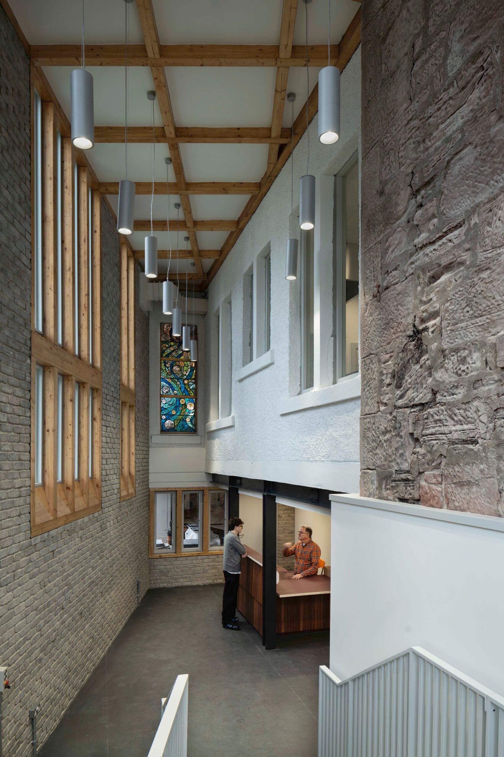 A hallway with brick and stone walls, large windows, a stained glass panel, and two people talking at a wooden reception desk. Ceiling lights hang from wooden beams above.