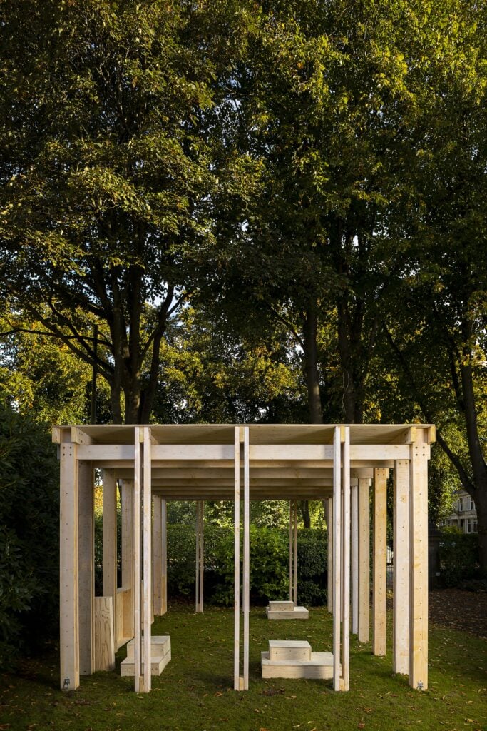 A rectangular wooden pavilion structure with open sides and benches stands on grass, surrounded by tall trees and greenery.