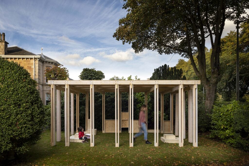 A wooden open-frame pavilion stands on a grassy lawn with one child seated and one adult walking inside; trees and a house are in the background.