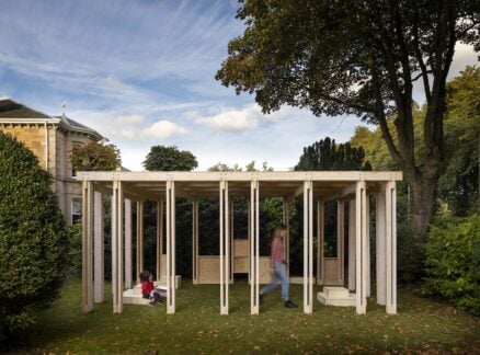 A wooden open-frame pavilion stands on a grassy lawn with one child seated and one adult walking inside; trees and a house are in the background.