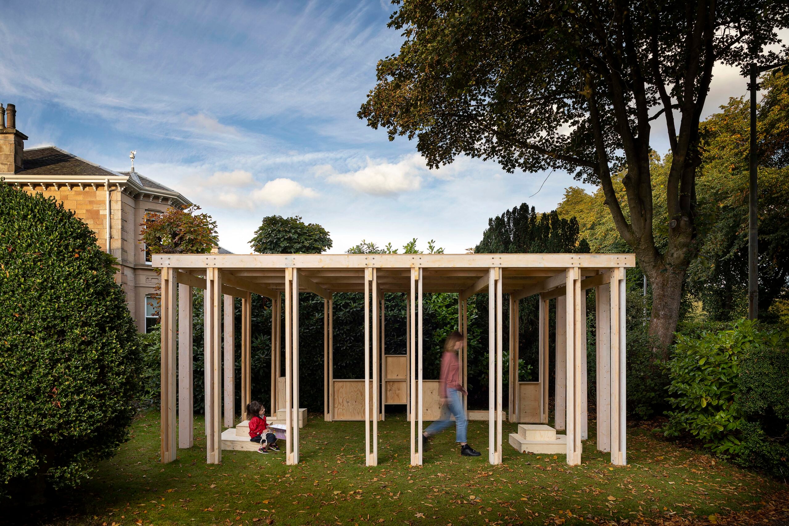 A wooden open-frame pavilion stands on a grassy lawn with one child seated and one adult walking inside; trees and a house are in the background.