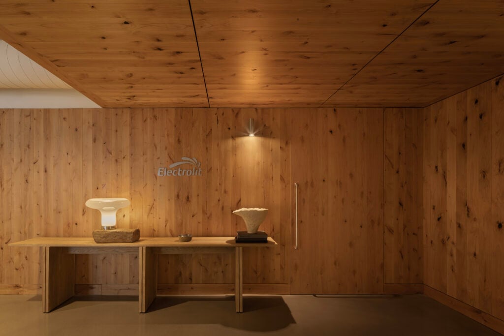 A minimalist wooden room features a BLDUS-designed bench with a decorative lamp, a bowl, and a sculptural object beneath an "Electrolit" logo on the wall.