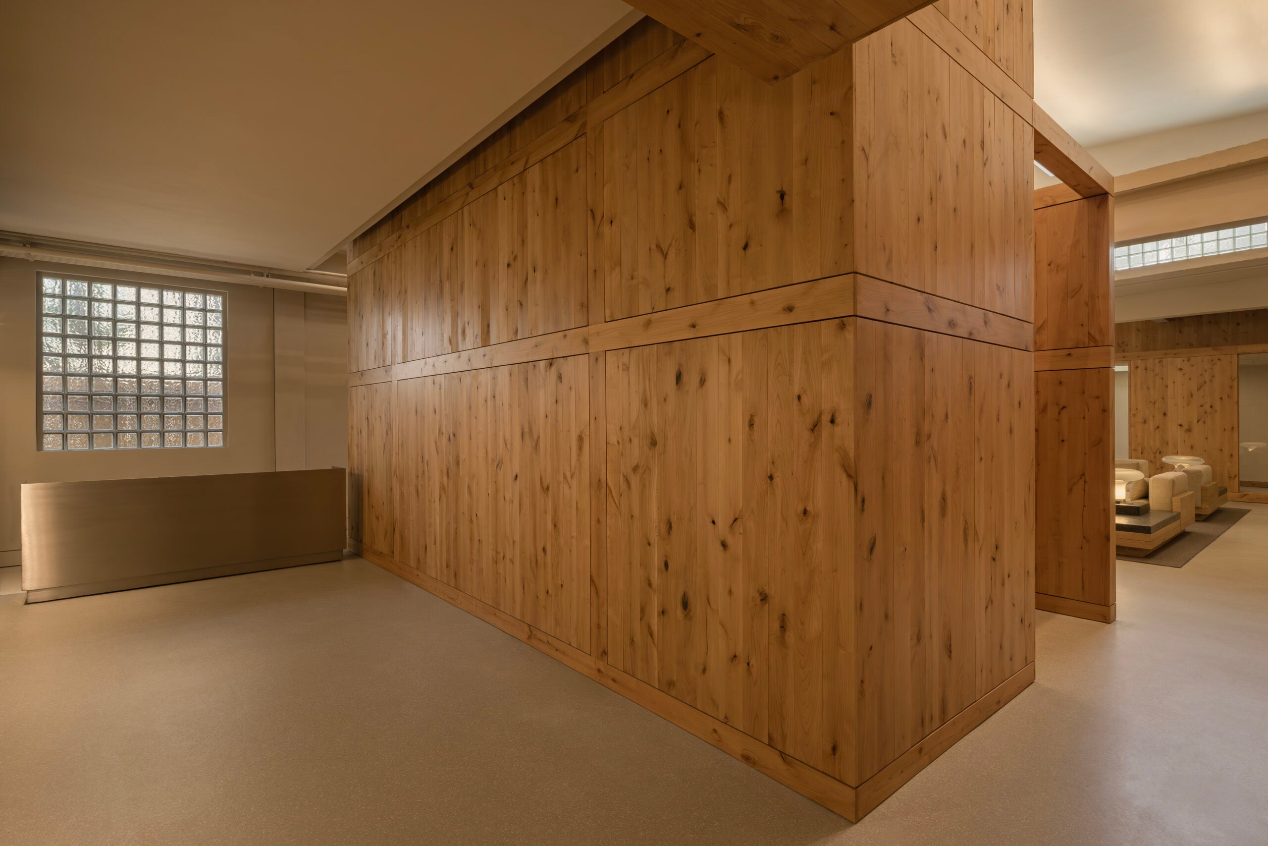 A modern interior space by BLDUS featuring large wooden wall panels, a frosted glass block window, beige flooring, and minimalist furnishings.