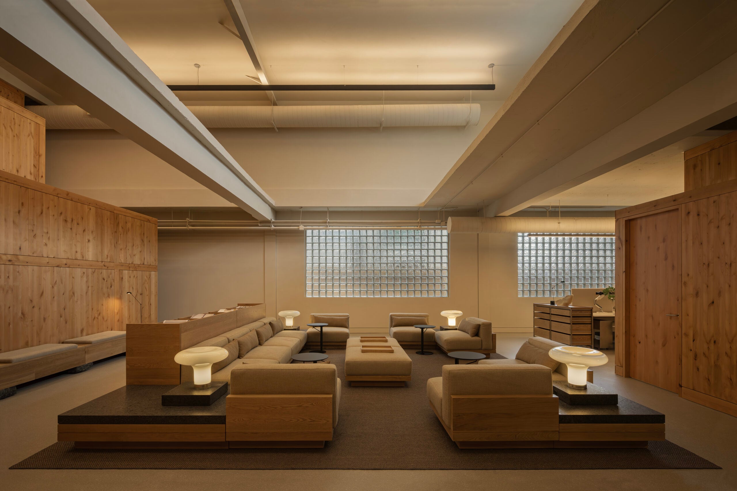 A spacious modern lounge by BLDUS features beige sectional sofas, wooden accents, cube lamps, and glass block windows under high ceilings with exposed beams.