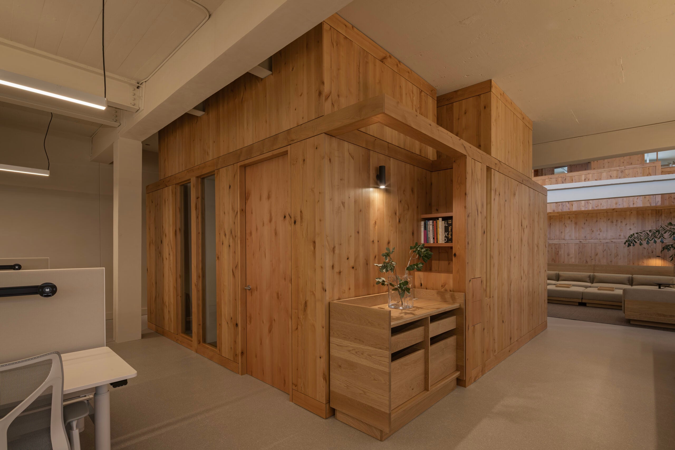 A modern office space featuring a BLDUS-designed wooden freestanding structure with a desk, shelves, and door, surrounded by open workspace and lounge area.
