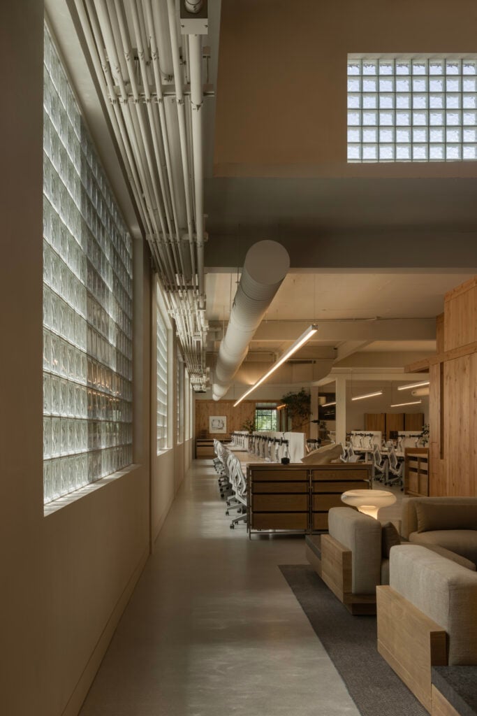 Modern office interior by BLDUS featuring glass block windows, exposed ceiling pipes, rows of white office chairs and desks, plus a cozy seating area with beige sofas and wooden partitions.