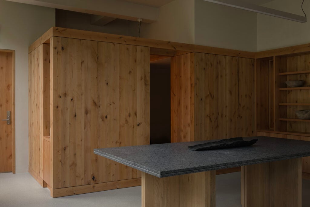 Interior view of a minimalist BLDUS kitchen with wooden cabinetry, a dark stone countertop island, and open shelving containing bowls.