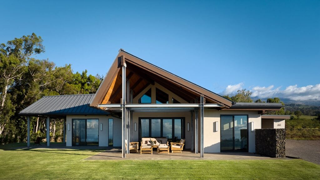 Modern Maui off-grid house with a steep gabled roof, large windows, and a covered patio with outdoor seating, set on a green lawn with trees and mountains in the background.