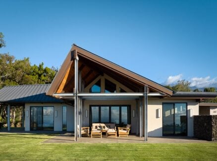 Modern Maui off-grid house with a steep gabled roof, large windows, and a covered patio with outdoor seating, set on a green lawn with trees and mountains in the background.