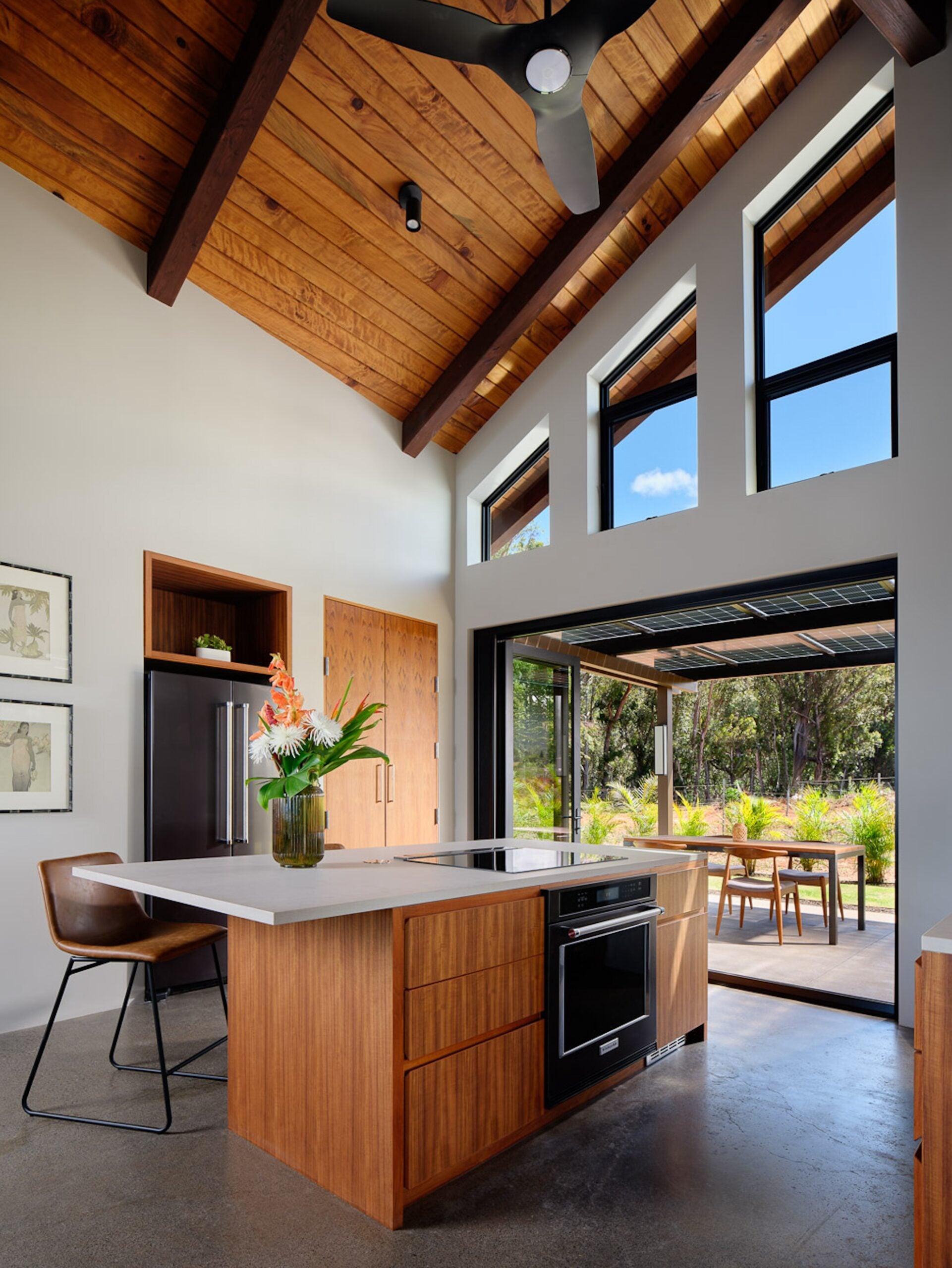 Maui Off-Grid House: Modern kitchen with wood island, high vaulted ceiling, large windows, and glass doors opening to an outdoor dining area; tropical flowers in a vase on the counter.