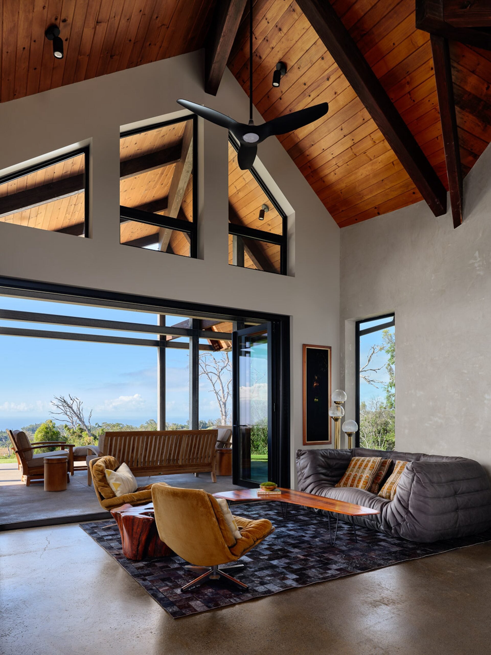 Modern living room with high wooden ceilings, large windows, and glass doors opening to an outdoor patio with seating and scenic views. Contemporary furniture and neutral tones are featured.