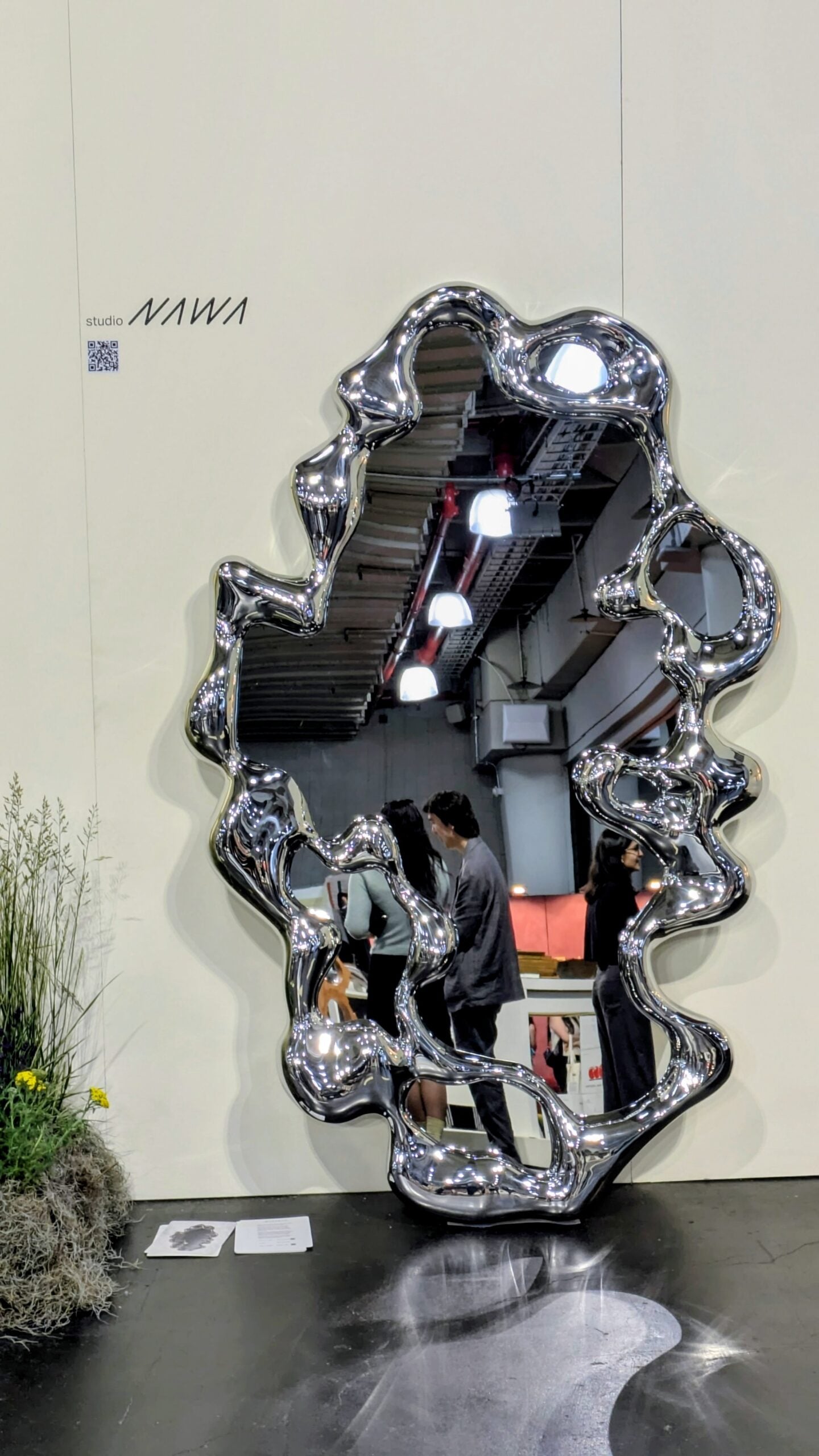 A large, irregularly shaped metallic mirror reflects people, ceiling lights, and intricate textiles at an indoor art exhibition. The mirror is mounted on a plain wall near a patch of grass and flowers.