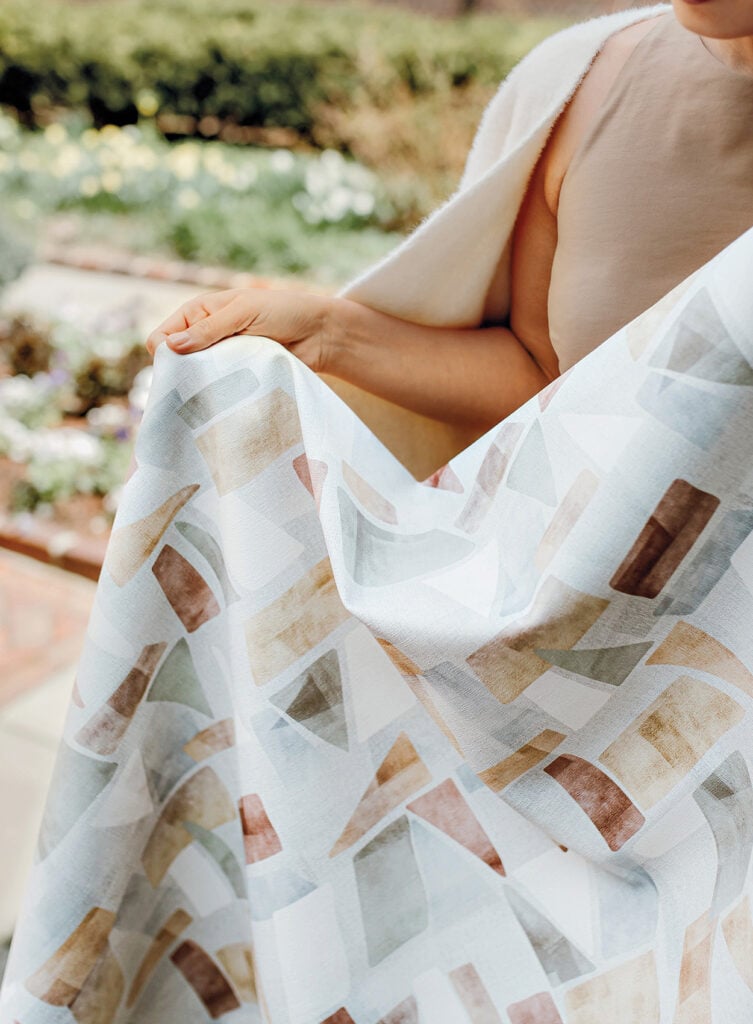 A person holds up a patterned material with geometric shapes in muted colors outdoors, with plants and flowers visible in the background.