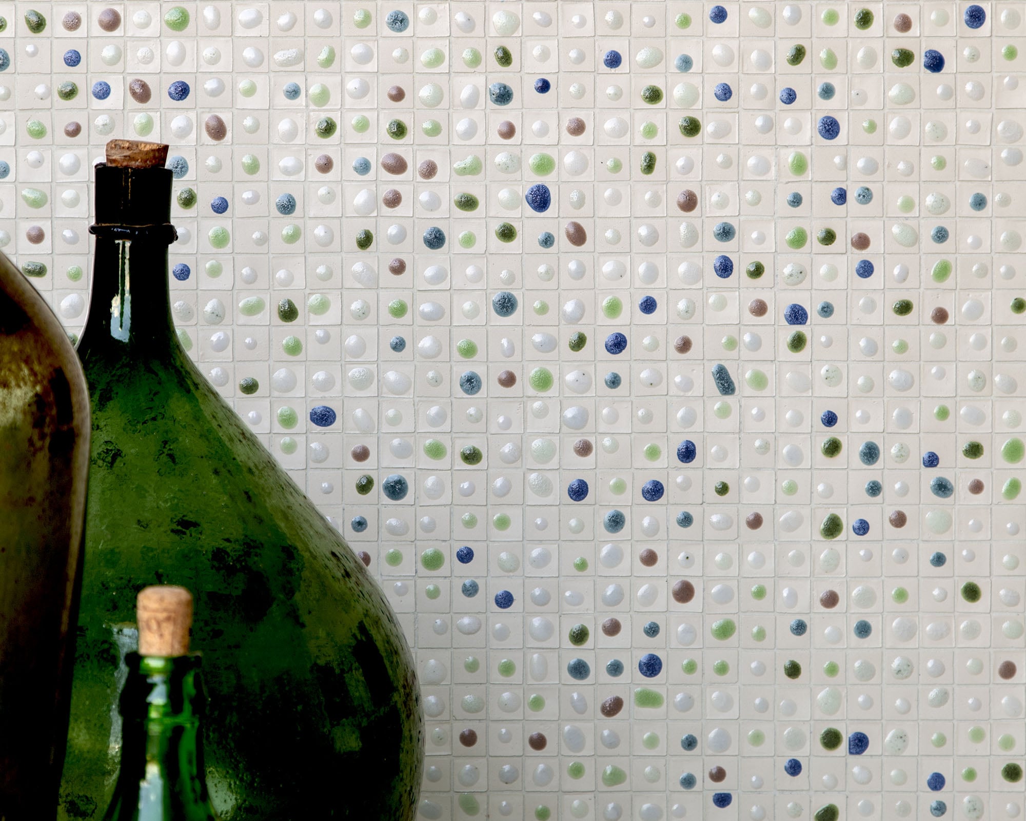 Two green glass bottles with cork stoppers are placed in front of a white tiled wall featuring small, colorful accents in blue, green, and brown—adding a touch of style reminiscent of artisanal textiles.