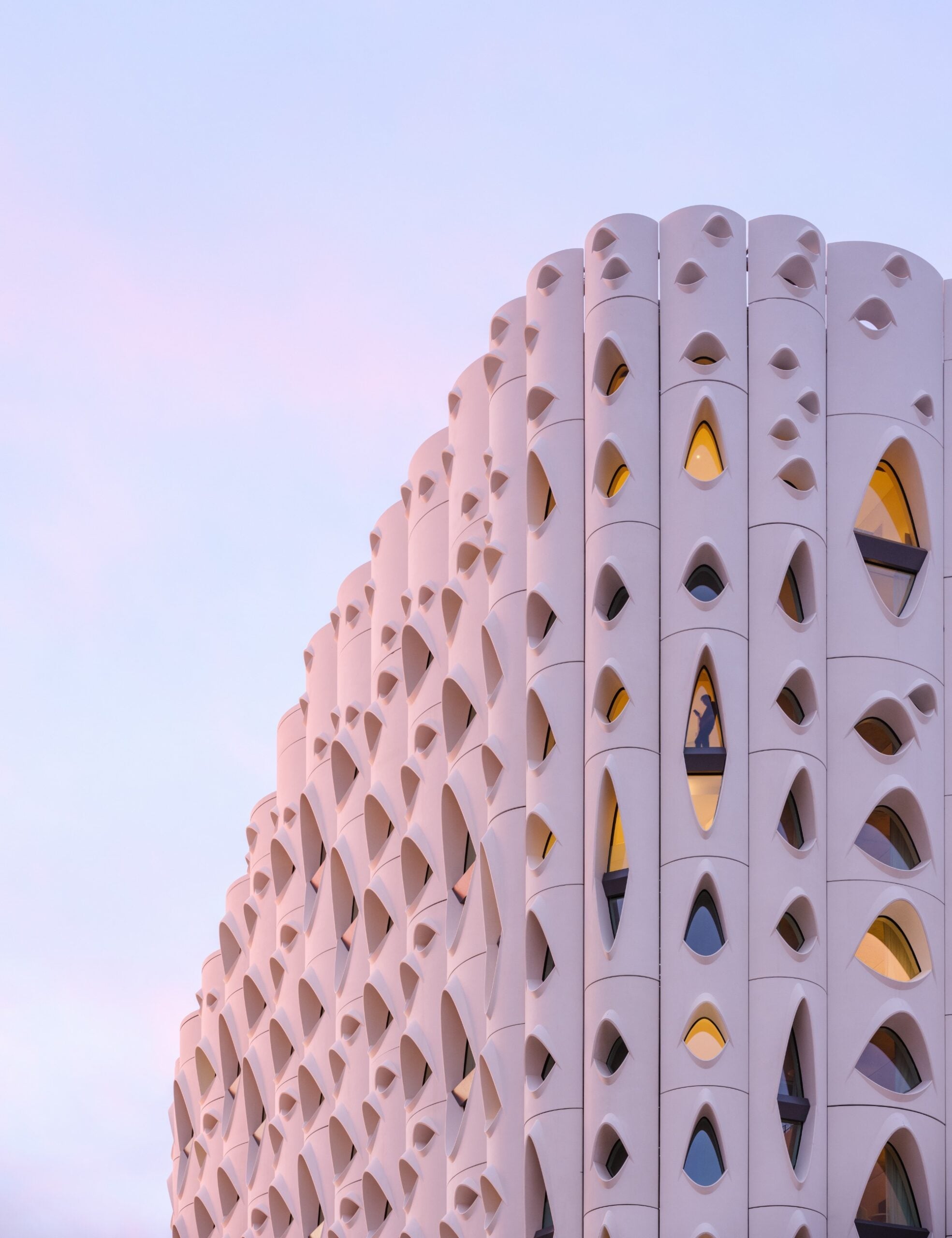 Modern building with a white, perforated facade featuring geometric cutouts and glowing windows at dusk, against a pastel sky—showcasing Wolf-Gordon’s innovative architectural materials.