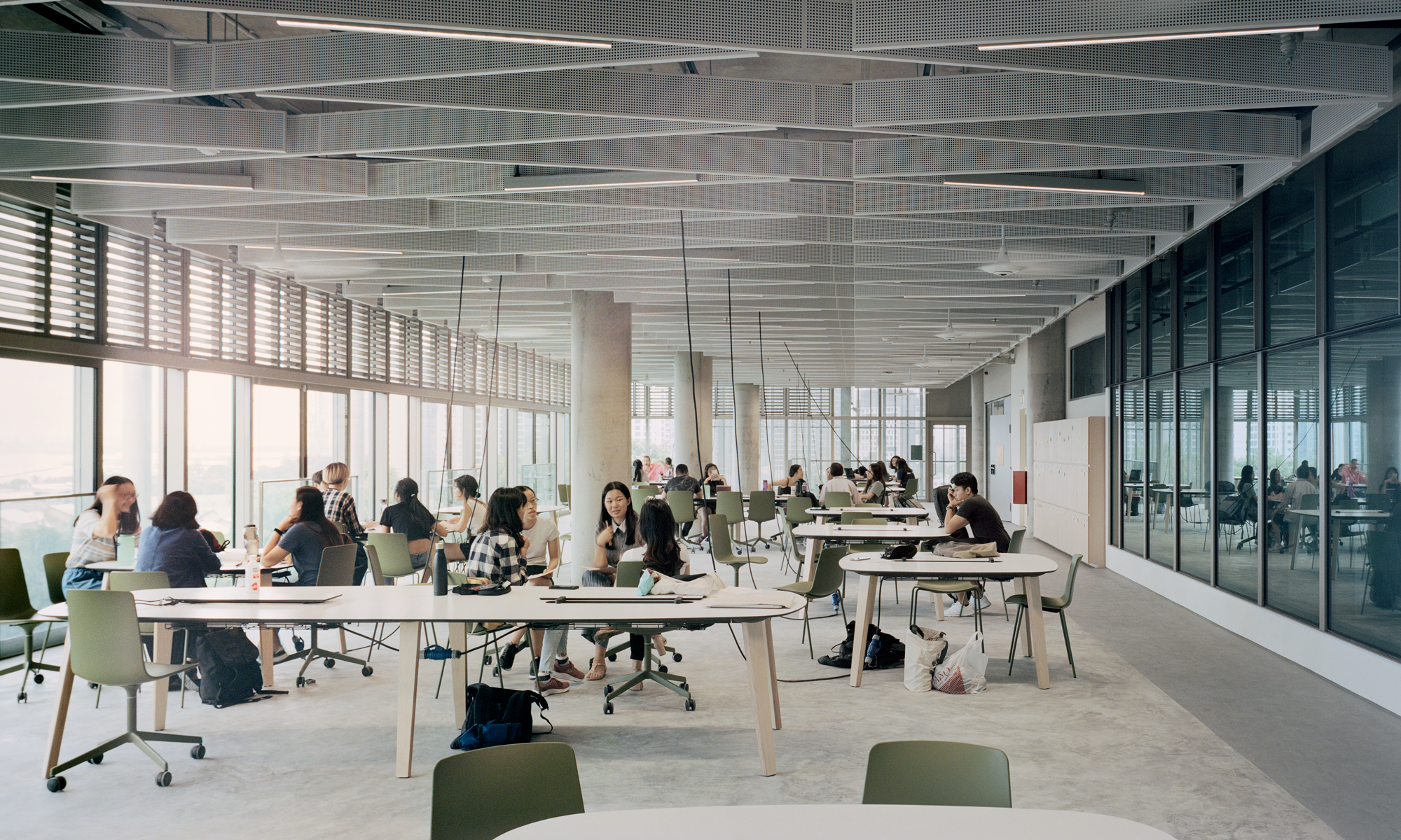 Modern open-plan workspace with people seated at tables, working and discussing key topics like Sustainability Roadblocks. Large windows provide natural light; glass wall and contemporary decor visible.