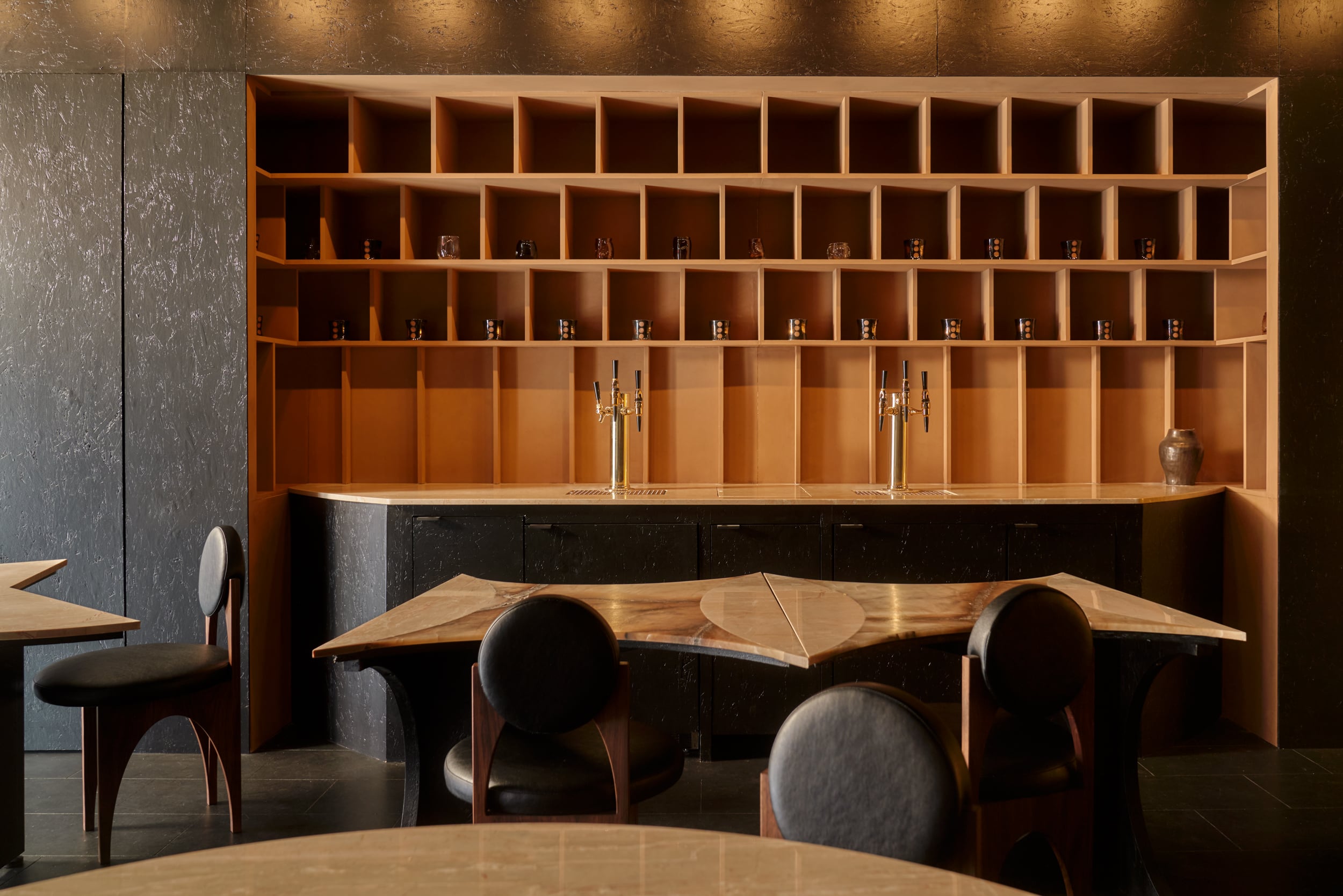 A modern, minimalist bar at Gourmega with empty shelves, three brass beer taps on a wooden counter, and several round tables with black chairs in the foreground.