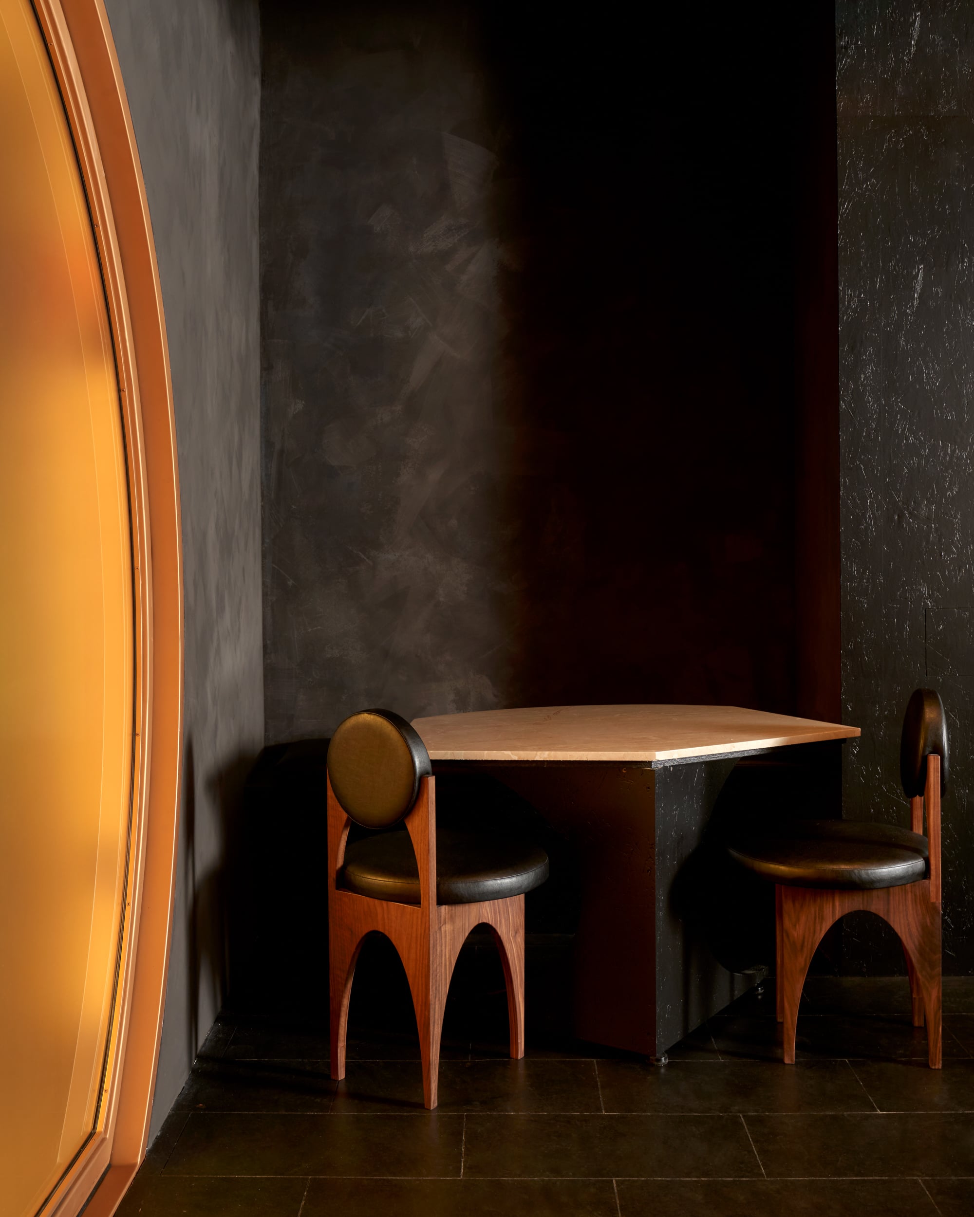 A small corner table at Gourmega restaurant with two wooden chairs in a dimly lit room with dark textured walls and a large amber-tinted window on the left.