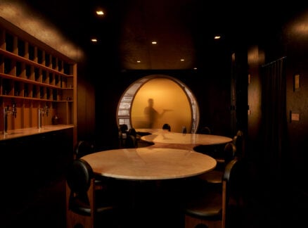 Dimly lit dining area at Gourmega with round tables and chairs; a frosted glass doorway reveals a blurred silhouette of a person pointing.