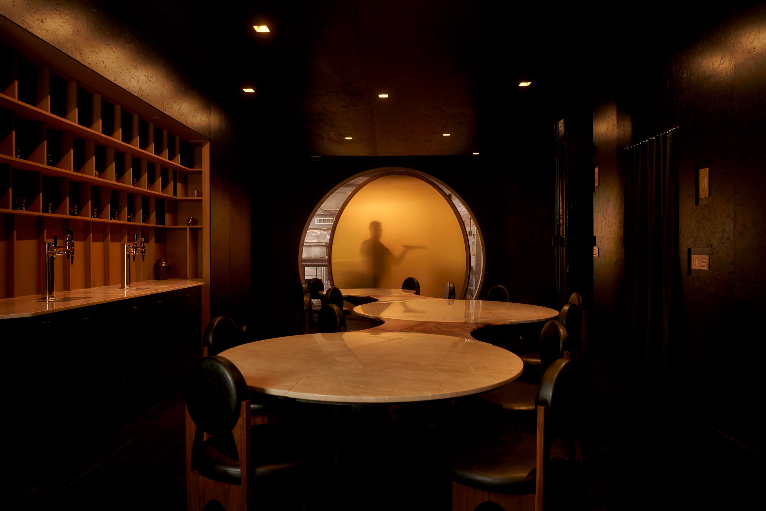 Dimly lit dining area at Gourmega with round tables and chairs; a frosted glass doorway reveals a blurred silhouette of a person pointing.