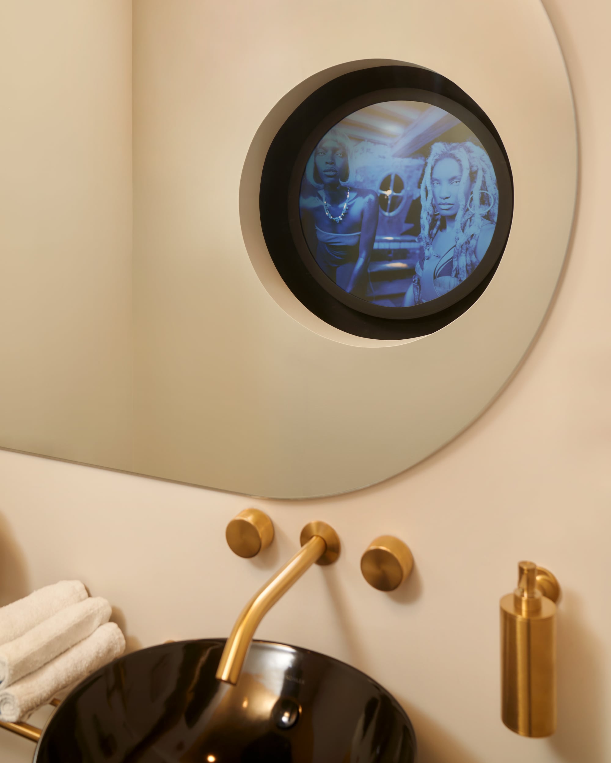 A round mirror above a bathroom sink displays a screen showing two people, with gold fixtures and towels visible nearby.