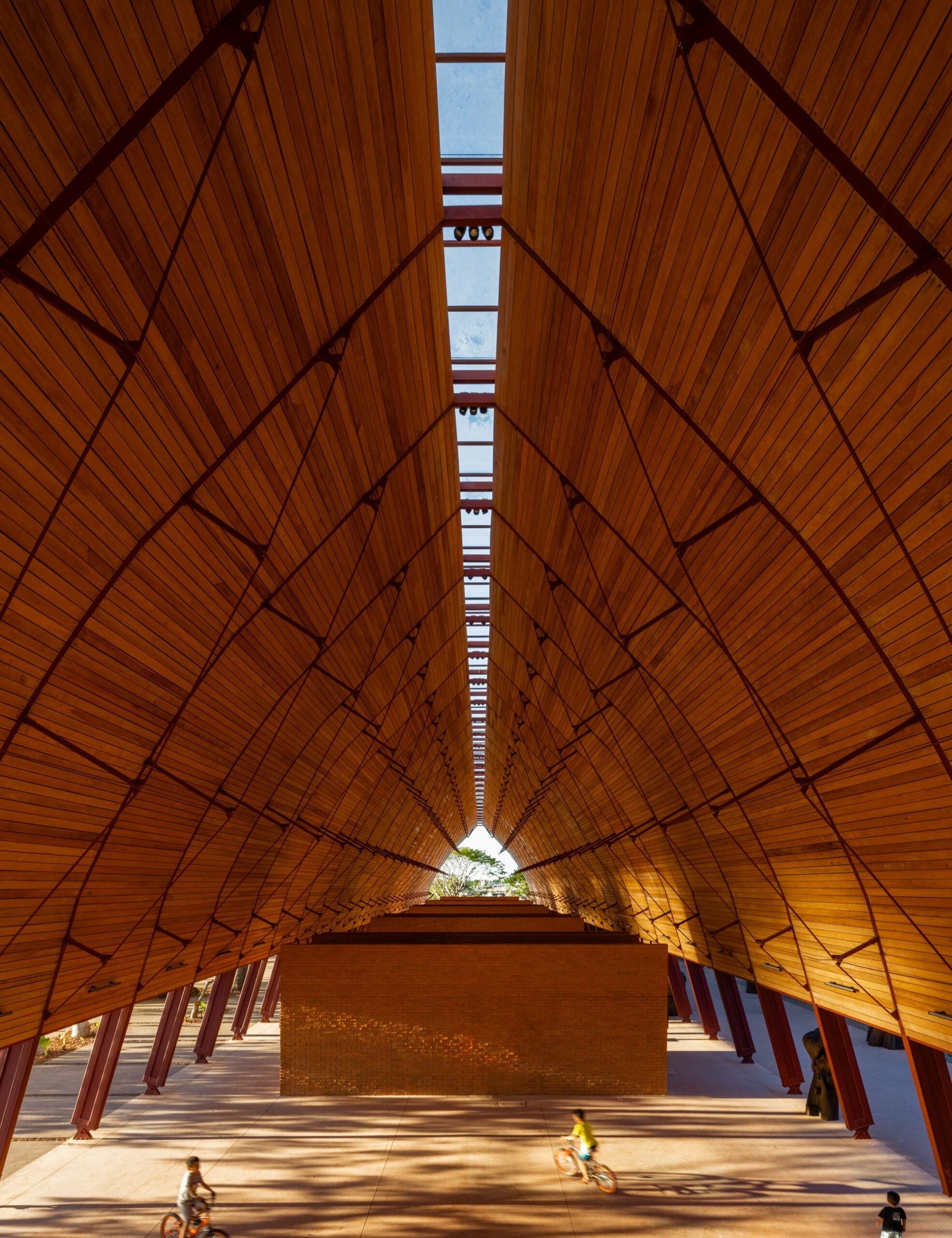 Symmetrical interior view of a large wooden pavilion with a high peaked ceiling, skylight, Wolf-Gordon finishes, brick wall at the end, and two cyclists passing through at ground level.
