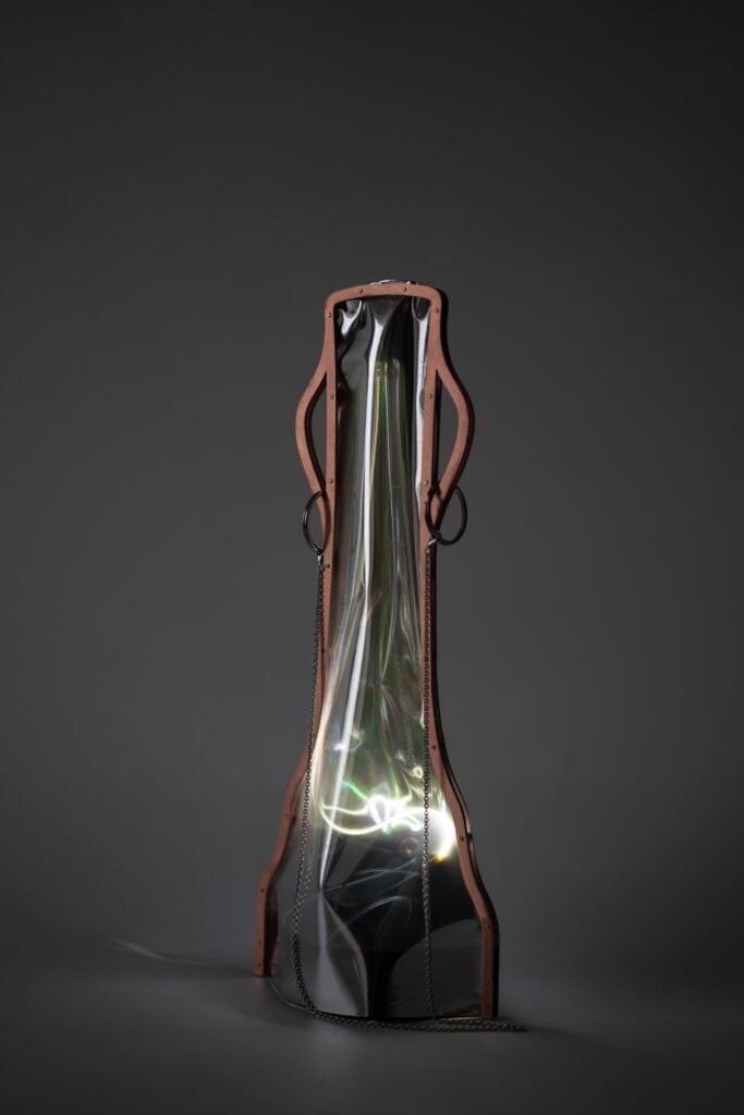 A sculptural object with a reflective, distorted metallic surface framed by brown material, chains, and rings, set against a dark gray background—an innovative piece inspired by Milan Design Week 2026.