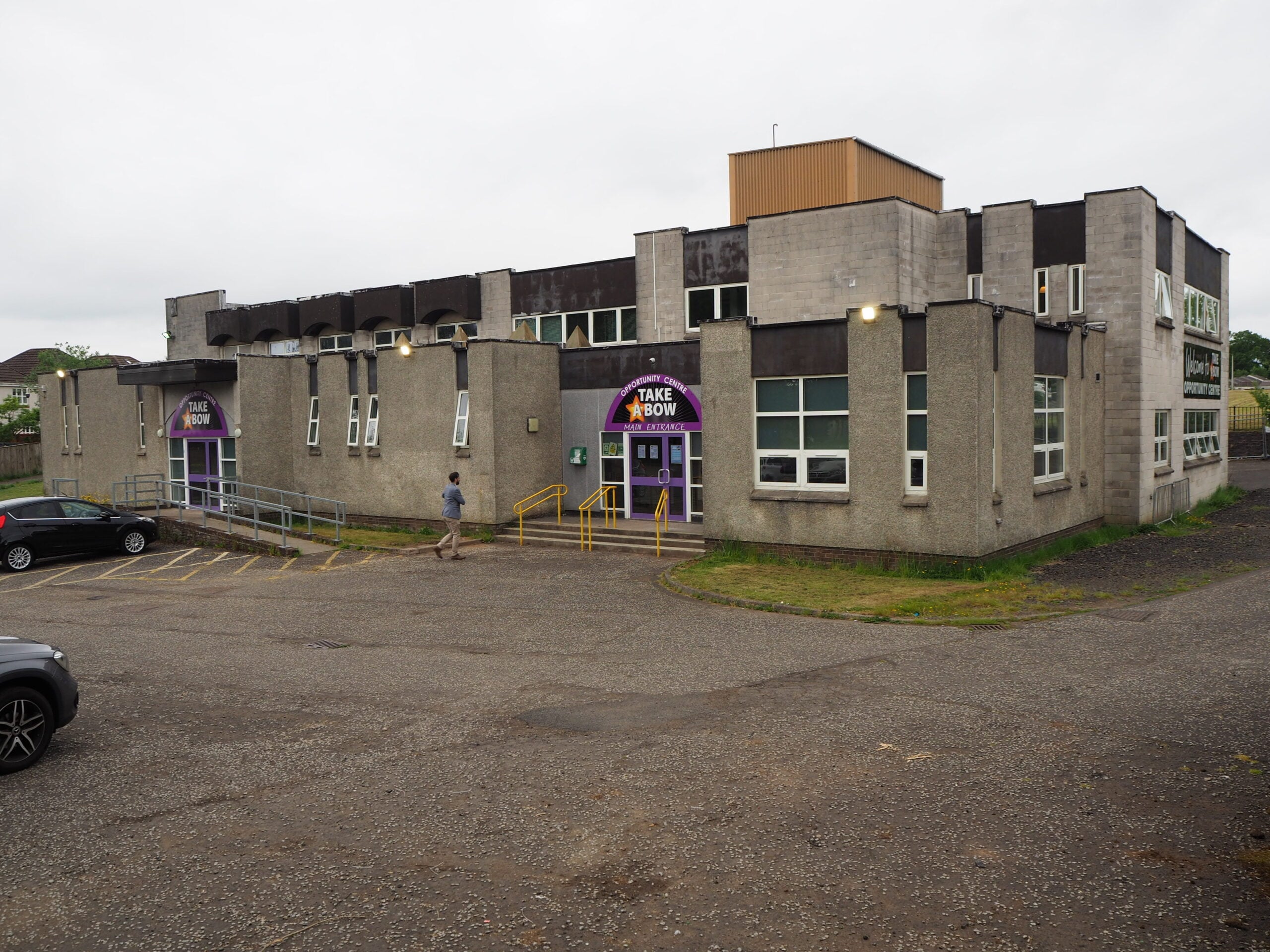 A two-story, grey concrete building with large windows and purple signs reading "Take A Bow," surrounded by a parking lot with several cars and a person walking toward the entrance.