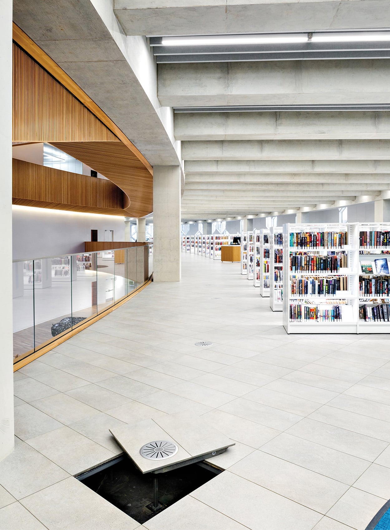 Modern library interior featuring rows of white bookshelves, an exposed concrete ceiling, and a floor access panel open to reveal venting material beneath.