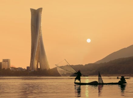Two people in a small boat fish with a net on calm water at sunset, with a modern twisted skyscraper and city skyline in the background—an image capturing the spirit of progress celebrated by the 2026 Vision Awards.