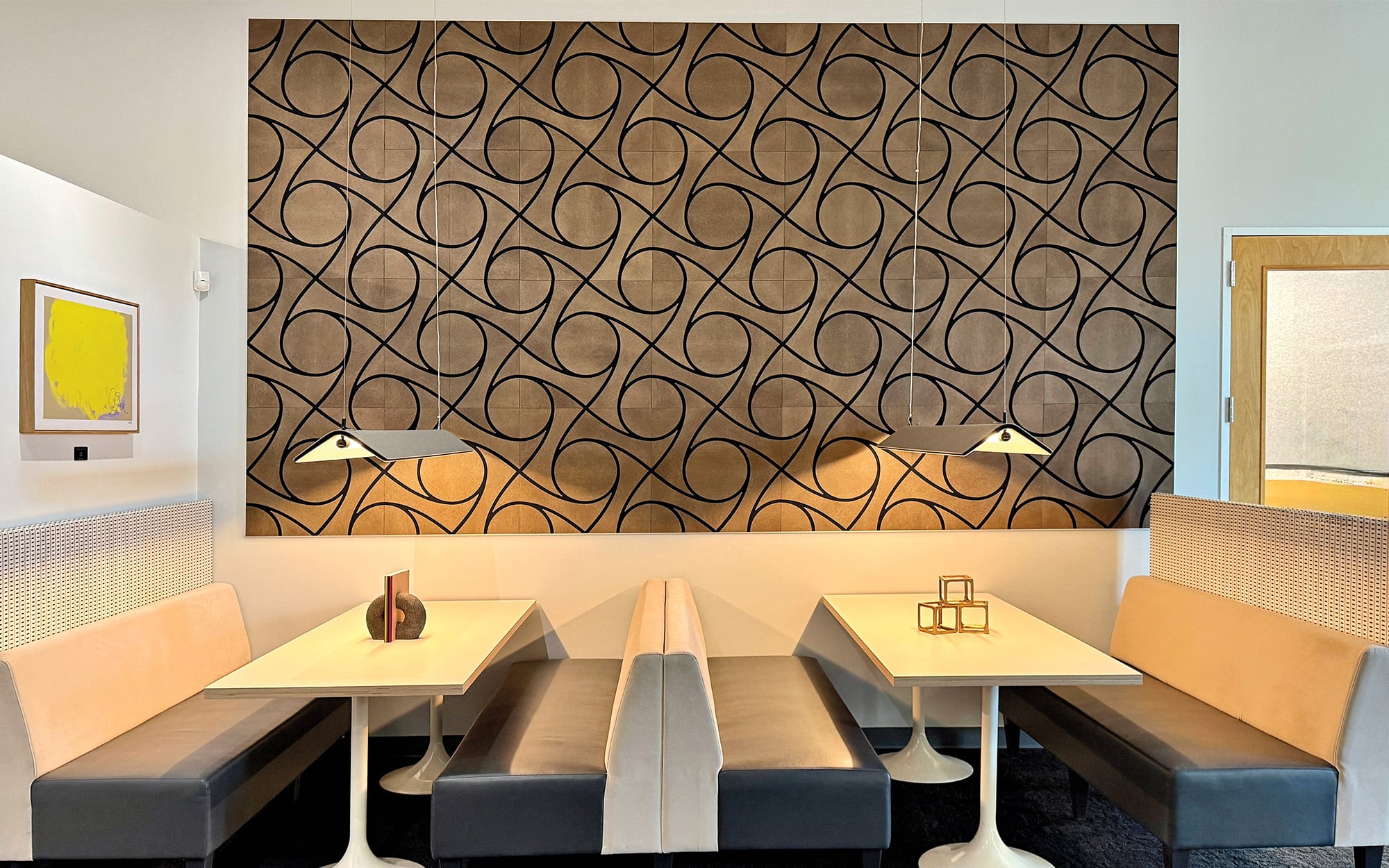 Two white tables with gray and beige booth seating are set against a wall with a geometric black swirl pattern, featuring sleek material finishes, two table lamps, and minimalist decor.