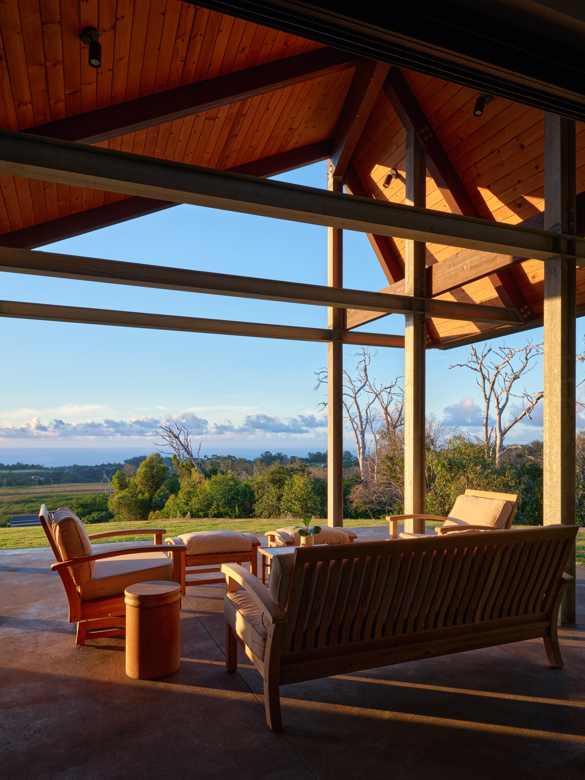 Wooden outdoor seating area under a pitched roof overlooks a scenic landscape with trees, open fields, and a partly cloudy sky at sunset.