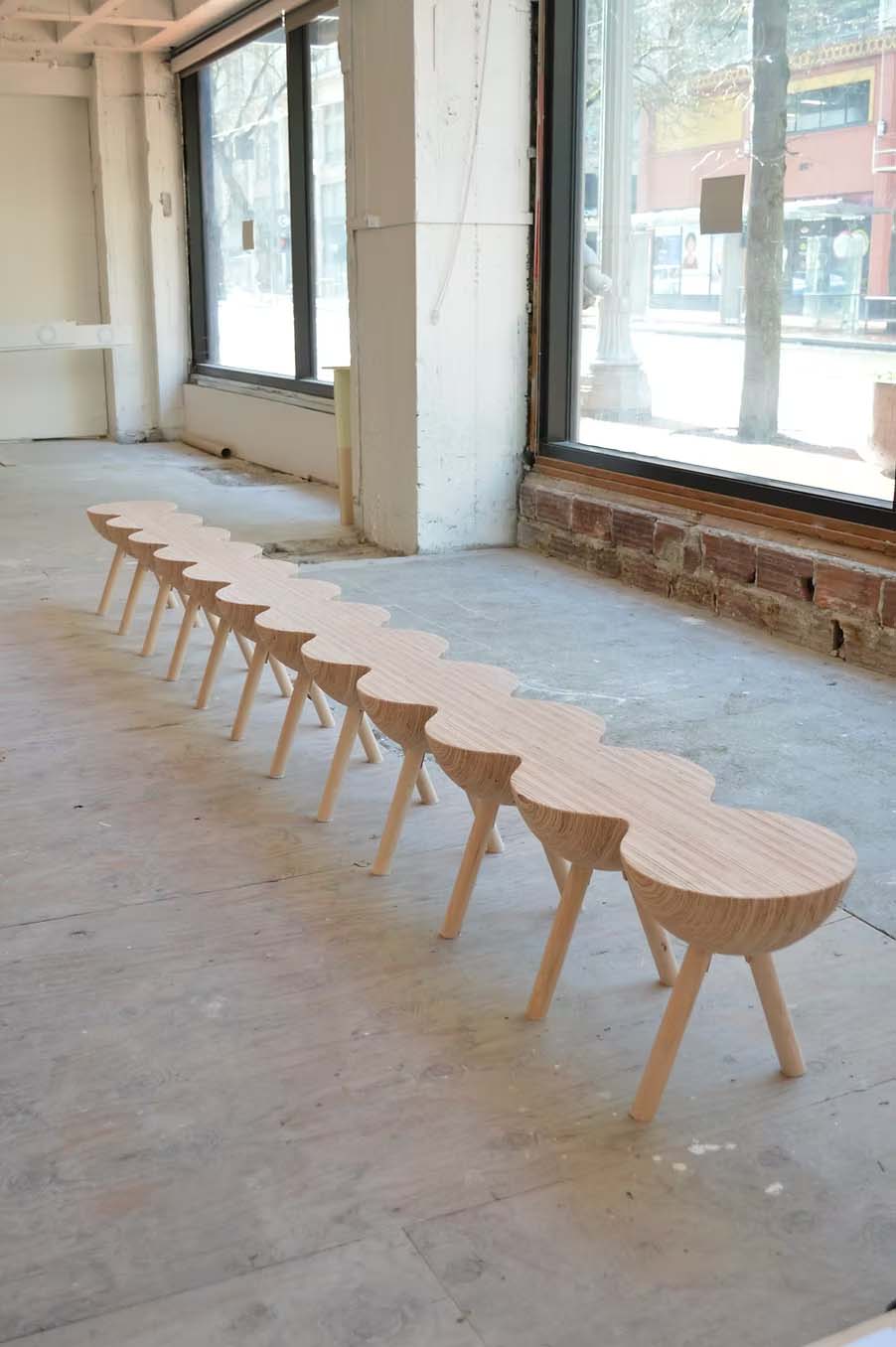 A long, light wood bench with a scalloped edge and angled legs sits in an empty, unfinished room with large windows, awaiting the addition of cozy textiles to soften its clean lines.
