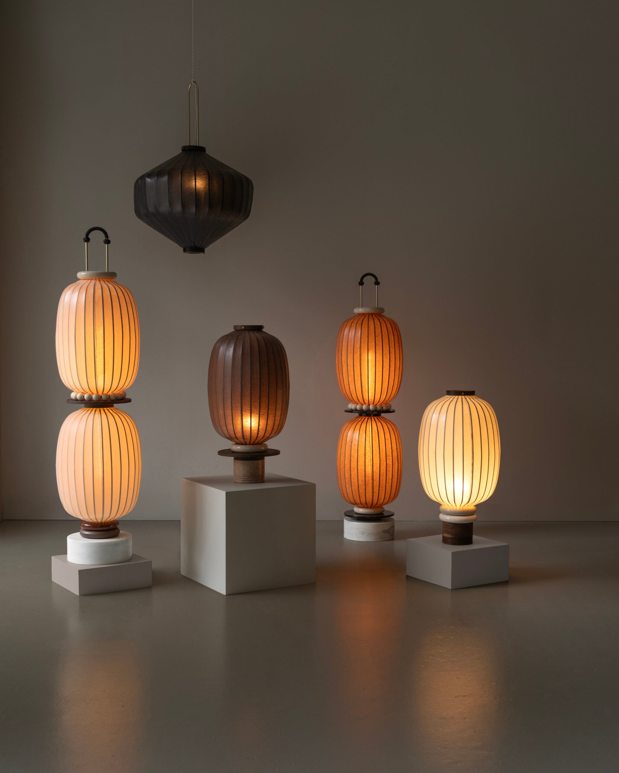 Five modern lantern-style lamps, three lit and two unlit, are arranged on and around white platforms in a minimal, neutral-toned room accented with soft textiles.