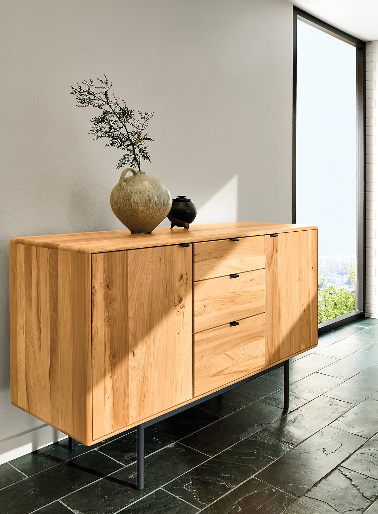 A wooden sideboard crafted from quality material with two cabinets and three drawers stands on metal legs; a ceramic vase with a branch and a small teapot sit atop, next to a large window.