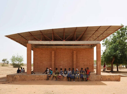 A group of people sit in the shade of a small brick building with a metal roof in a dry, rural area with sparse trees, evoking scenes discussed in MOS’s New Book On Public Spaces.